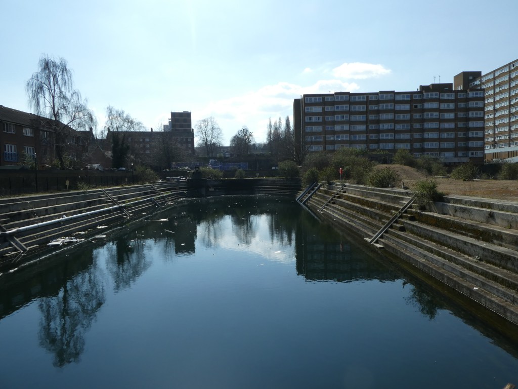 A photograph of a former dock, filled with water, with a small block of flats at the rear left and a larger one at rear right.