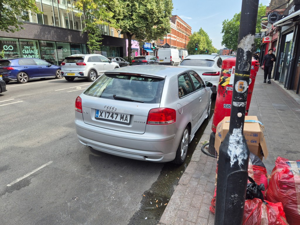 A silver car with the number plate "X 1747 MA", in a parking bay next to some rubbish bags and cardboard boxes.