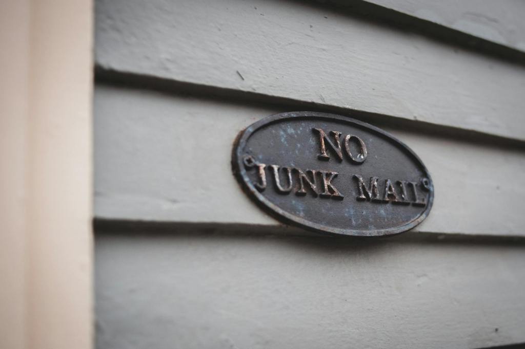 A metal sign on a wooden fence saying "No Junk Mail" in serif capital letters", somewhat rusted.